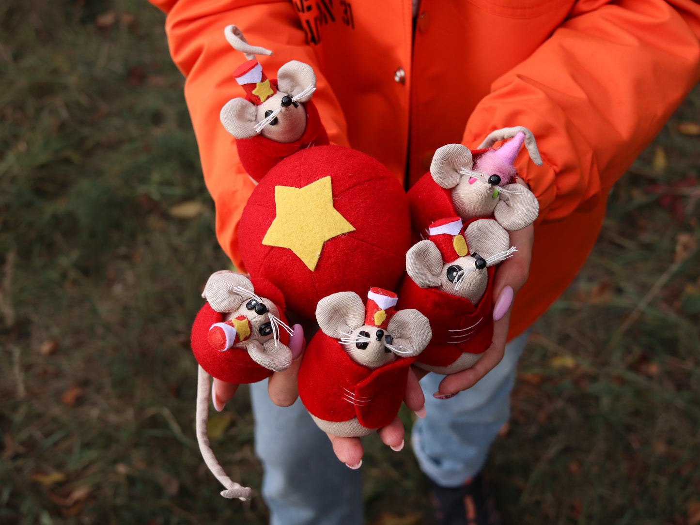 Person holding red plush toys shaped like mice with a yellow star, wearing an orange jacket.