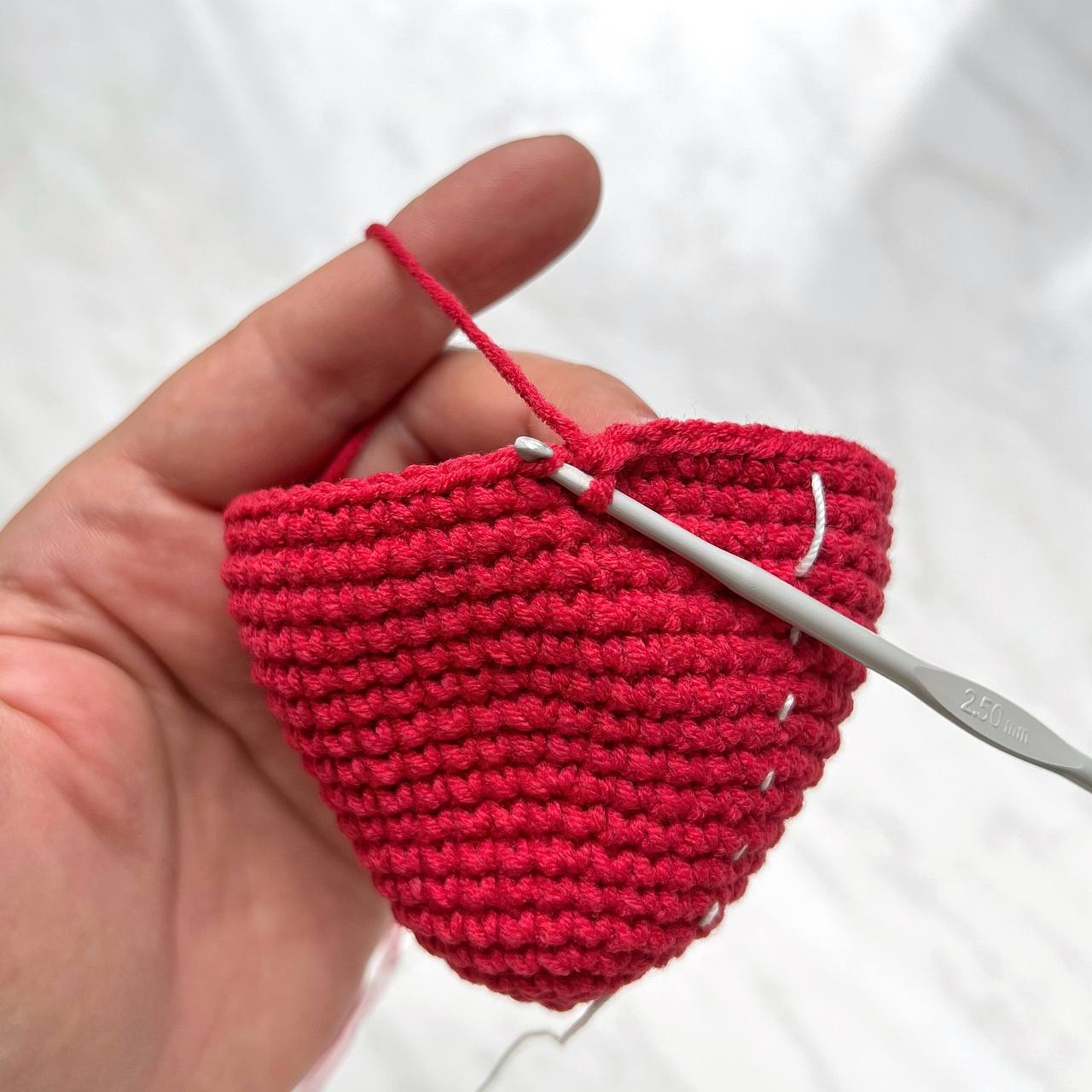 Hand holding a red crochet heart with a crochet hook on a white background