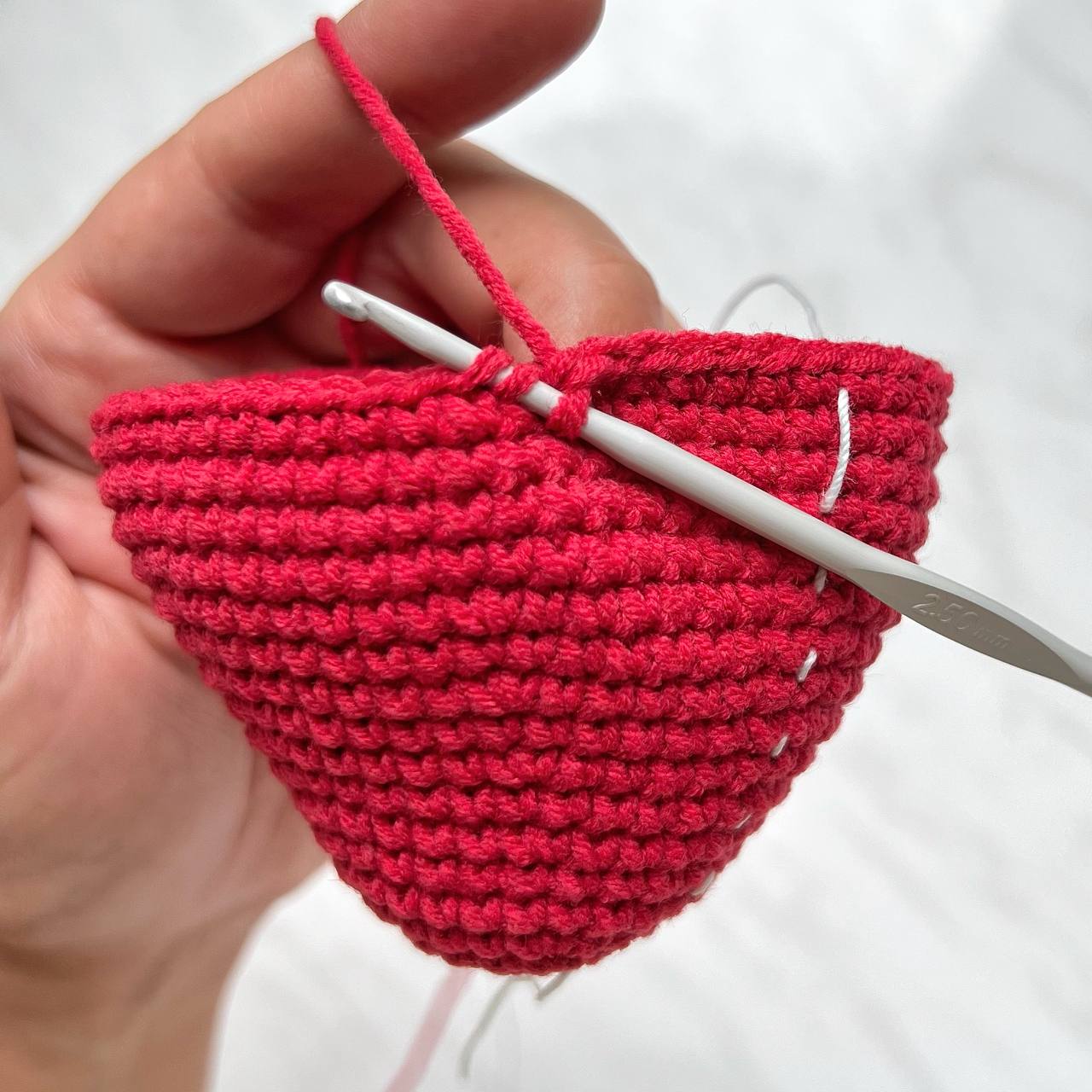 Hand holding a red crochet heart with a white crochet hook against a white background