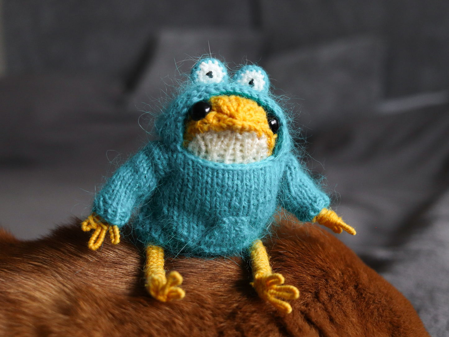 Knitted toy resembling a frog with a blue body and yellow feet and beak, sitting on a brown surface.