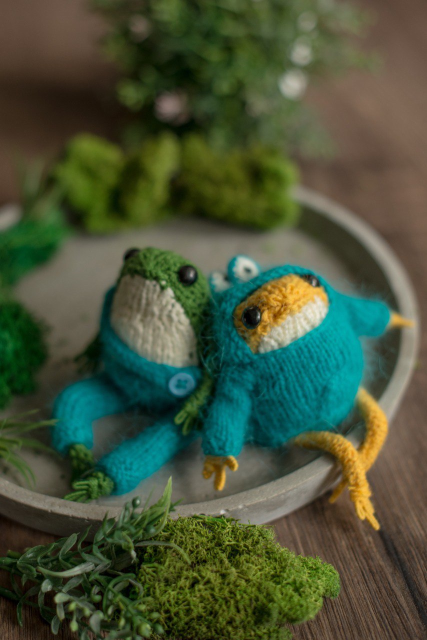 Knitted frog toys on a round wooden surface with greenery.