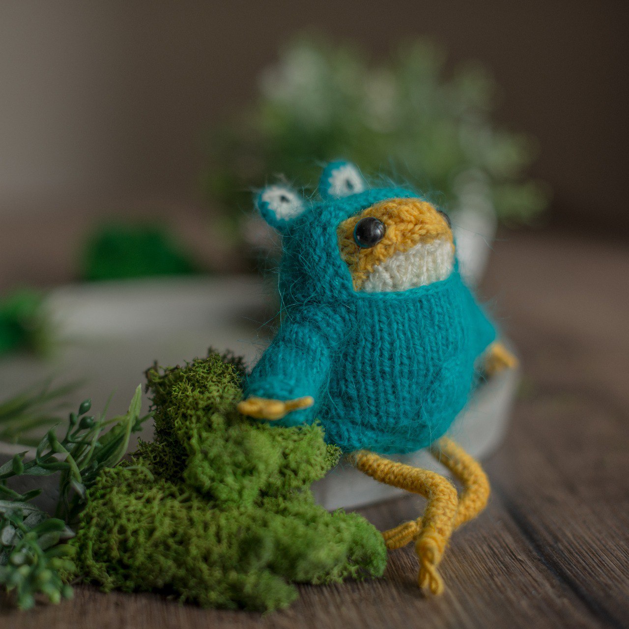 Knitted blue frog in a sweater with yellow accents on a wooden surface with greenery