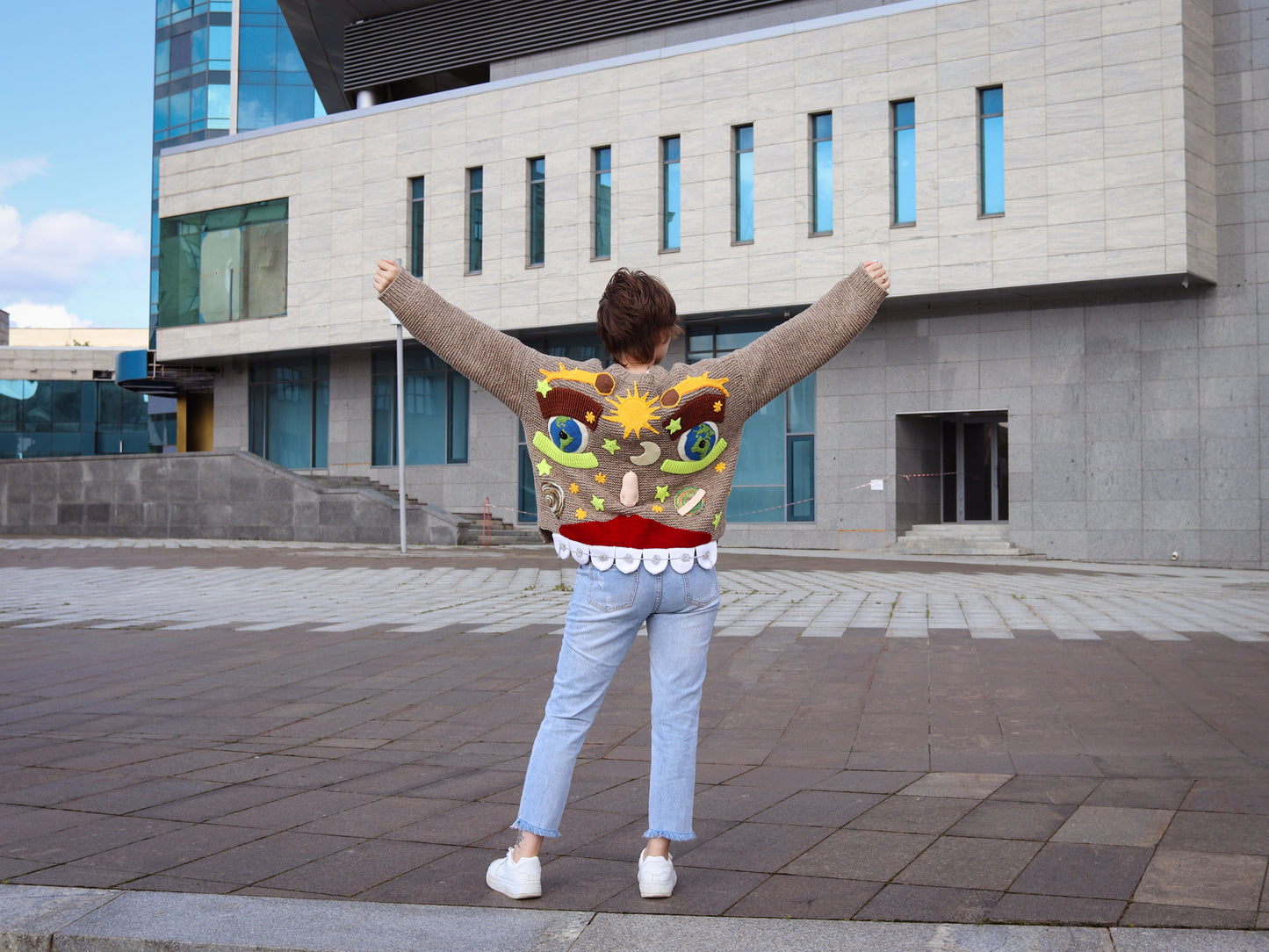 Person wearing a colorful sweater with a cartoon design, standing in front of a modern building.