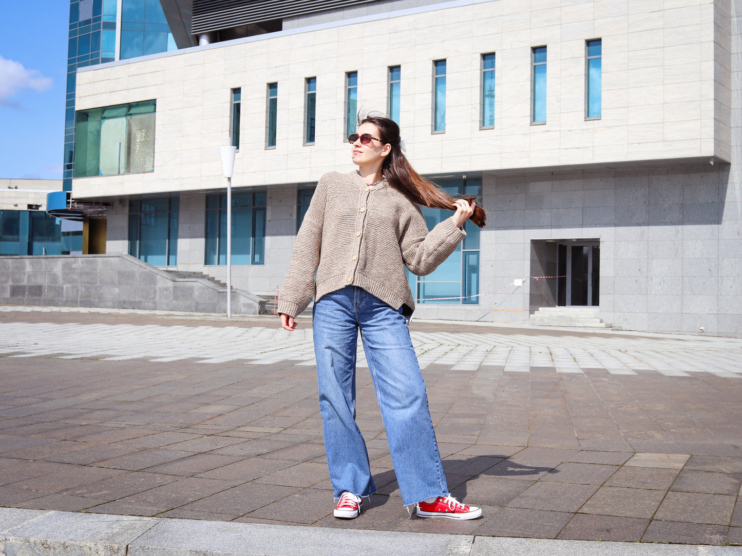 Woman in a beige cardigan and blue jeans standing on a sidewalk with a modern building in the background