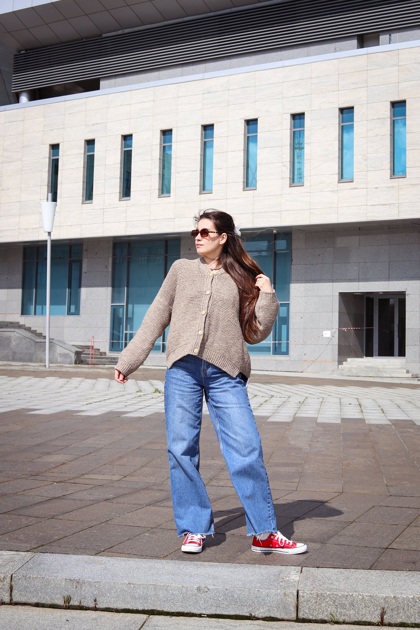Woman standing outdoors in front of a building wearing a beige cardigan, blue jeans, and red sneakers.