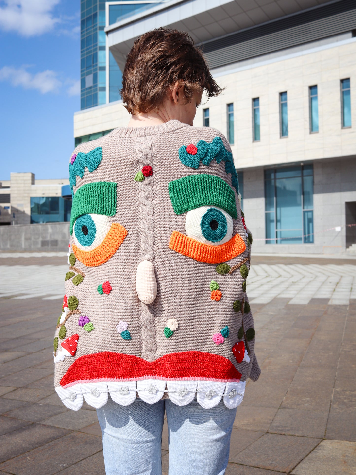 Person wearing a colorful, cartoonish cardigan with large eyes and a red mouth outdoors.
