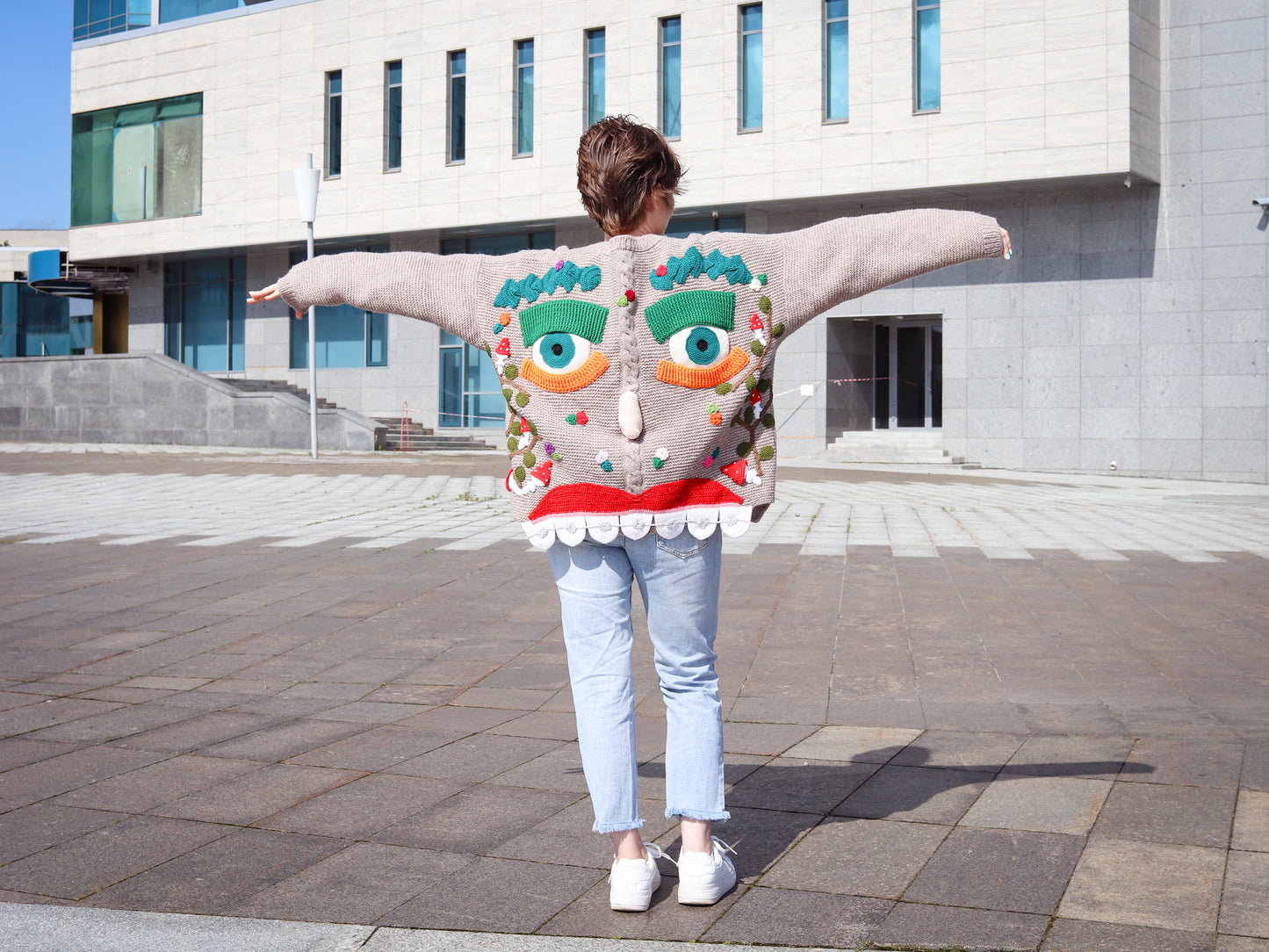 Person wearing a colorful, mask-like sweater with a modern building in the background