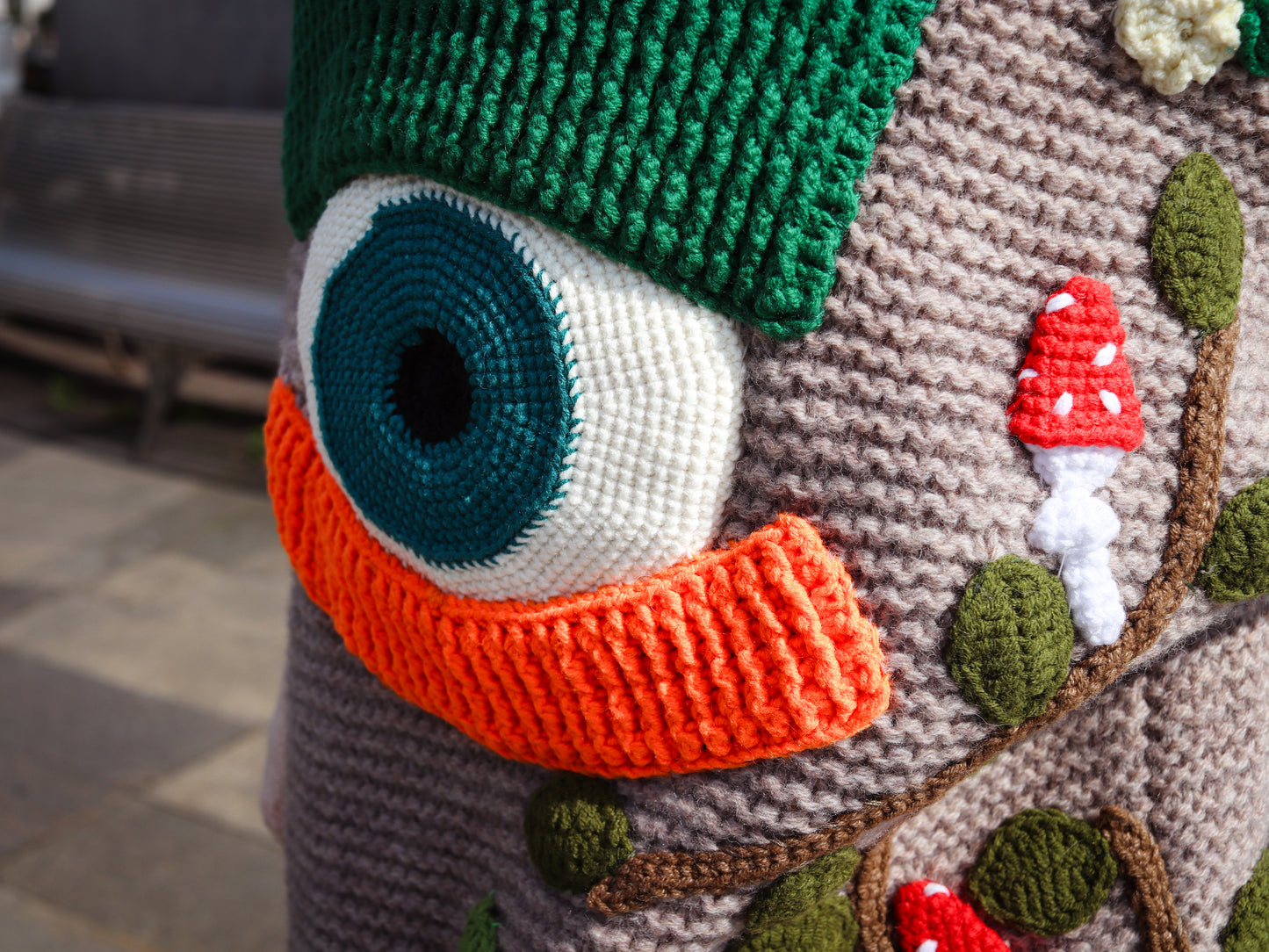 Close-up of a crocheted creature with large eye, orange beak, and mushroom decorations.