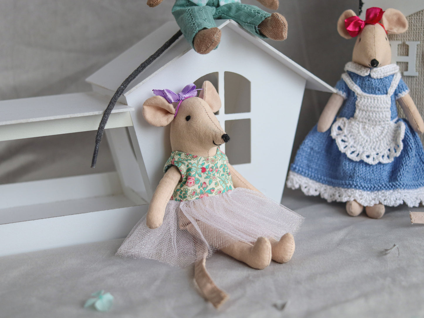 Two plush mouse toys in dresses sitting on a surface with a small house-like structure in the background.