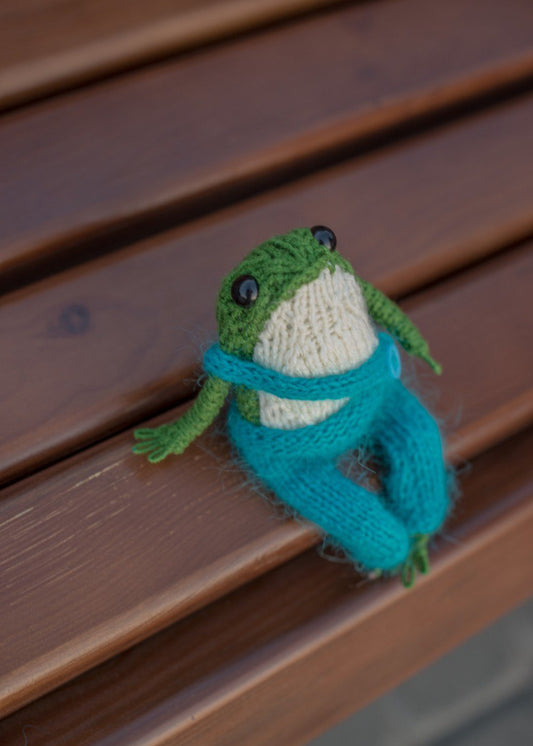 Plush frog toy in knitted sweater sitting on wooden surface